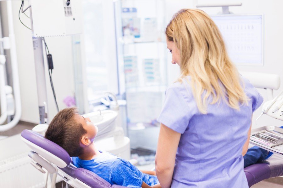 Young boy sitting in a dental chair while a female dental professional attends to him in a bright dental clinic.